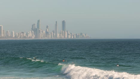 Surfers-enjoying-the-waves-on-a-sunny-day,-Burleigh-Heads-with-Surfers-Paradise-in-the-background,-Gold-Coast,-Australia