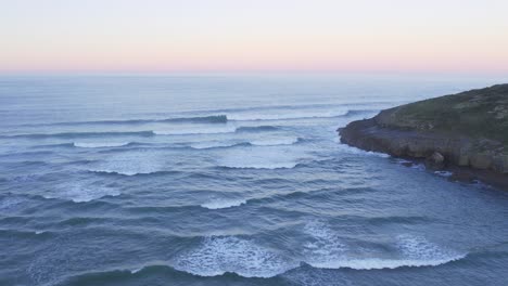 Sanfte-Wellen-Entlang-Der-Zerklüfteten-Küste-Bei-Sonnenuntergang-Schaffen-Eine-Malerische-Szene-Aus-Der-Luft-In-4K