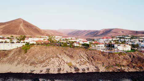 Ciudad-Costera-En-Fuerteventura-Con-Paisaje-Volcánico-Y-Cielos-Despejados,-Vista-Aérea