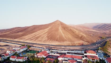 Quaint-town-nestled-at-the-base-of-a-majestic-mountain-in-Fuerteventura,-clear-skies,-aerial-view