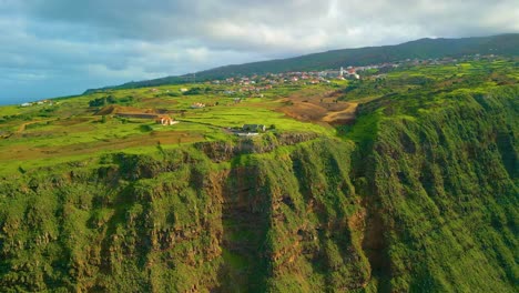 Drone-shot-showing-gigantic-steep-greened-cliff-of-Madeira-and-small-village-on-top-of-mountain---approaching-drone-shot