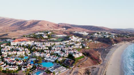 Coastal-resort-in-Fuerteventura-with-white-buildings-and-pools,-flanked-by-hills-and-a-beach