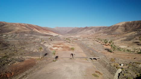 Paisaje-árido-Con-Autopista-En-Fuerteventura,-Escasa-Vegetación-Bajo-Un-Cielo-Azul-Claro.