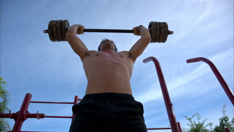 Slow-motion-tilt-up-shot-of-an-hispanic-shirtless-man-performing-several-barbell-front-squat-to-overhead-press-at-the-fitness-ground-of-the-park