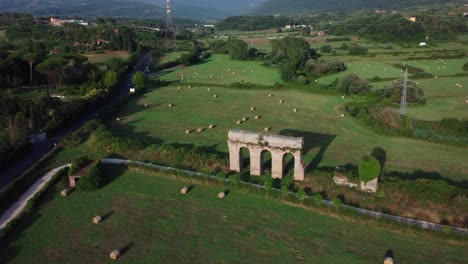Ancient-Roman-aqueduct-ruins-in-pastoral-Lazio-landscape,-Italy,-hay-bales-scattered-in-fields,-aerial-view