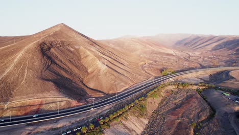 Kurvenreiche-Straße-Durch-Karge-Hügel-Auf-Fuerteventura,-Mit-Spärlicher-Vegetation-Und-Geparkten-Autos,-Luftaufnahme