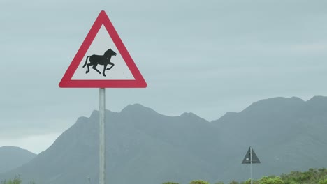 Triangular-caution-road-sign-with-a-horse-on-it-and-huge-mountains-in-the-background