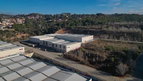 Industrial-warehouses-near-wooded-area-in-Abrera,-Barcelona,-Spain,-under-clear-skies,-aerial-view