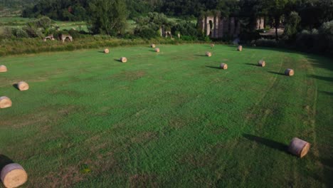Hay-bales-on-green-field-near-ancient-Roman-Aqueduct-in-Montalto,-Lazio,-Italy,-sunny-day,-aerial-view