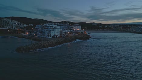 Twilight-descends-on-Roc-Sant-Gaietà,-coastal-village-in-Tarragona,-Spain,-lights-glisten,-waves-gently-break