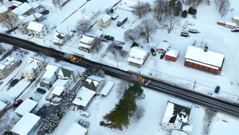 Two-snowplow-trucks-with-blades-and-salt-driving-on-road-through-USA-town-covered-in-winter-snow