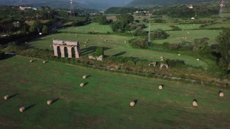 Ancient-Roman-Aqueduct-in-Lazio,-Italy,-surrounded-by-green-fields-and-hay-bales-in-the-golden-light,-aerial-view