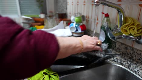 Hands-of-old-woman-cleaning-grey-baking-tray-in-kitchen