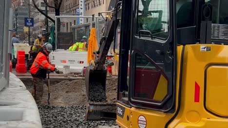 A-low-angle-shot-of-a-man-shoveling-gravel-on-a-construction-site-in-Manhattan,-New-York-with-another-man-in-an-excavator-assisting-him