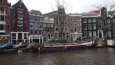Blick-Auf-Das-Stadtbild-Von-Amsterdam-Mit-Holländischen-Architekturgebäuden,-Herumlaufenden-Fußgängern-Und-Hausboot