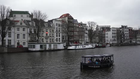 Blick-Auf-Das-Stadtbild-Von-Amsterdam-Mit-Einem-Transportboot,-Das-Durch-Den-Kanal-Fährt,-Malerische-Häuser-Im-Holländischen-Architekturstil-Im-Hintergrund