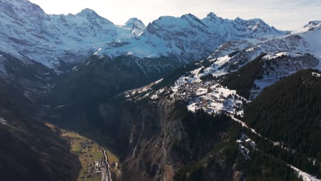 Scenic-aerial-view-of-Switzerland-village-near-steep-mountain-edge,-snowy-peaks