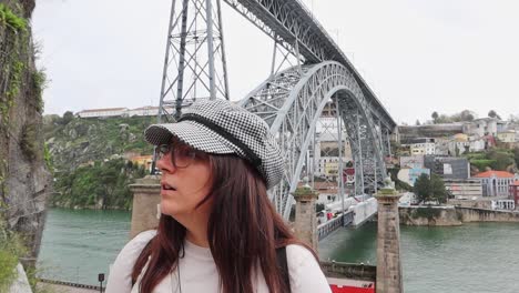Stylish-woman-in-glasses-and-checkered-hat-stands-in-front-of-Ponte-Luis-I,-Porto