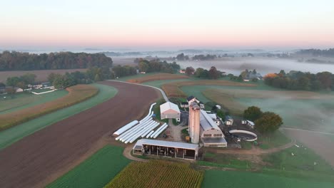 Aerial-dawn-view-of-a-farm-with-silos,-multiple-barns,-patchwork-fields,-a-house,-and-fog