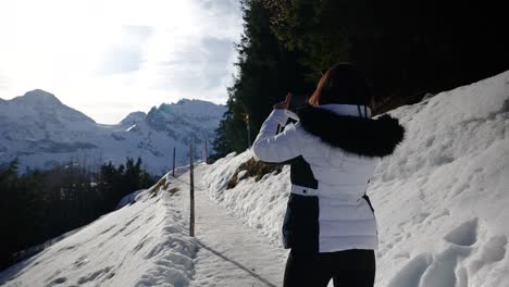 Female-tourist-stand-on-snowy-Switzerland-mountain-trail-and-take-photos