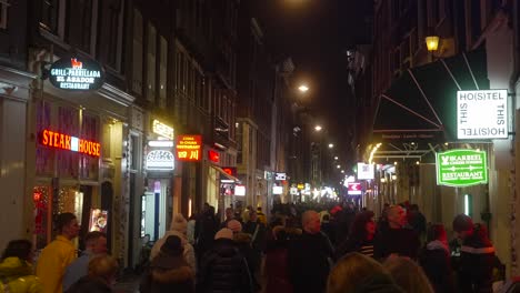 People-walking-around-busy-city-center-street-in-Amsterdam,-shops,-restaurants,-hotels-night-time