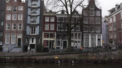 Pedestrians-walking-along-road-next-to-the-canal-and-dutch-architectural-building-in-downtown-Amsterdam