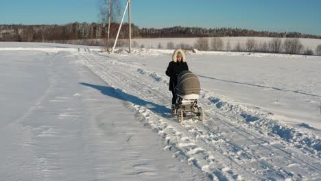 Mother-in-dark-winter-jacket-walk-with-baby-carriage-on-snowy-countryside-road