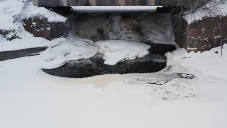 Dark-water-stream-from-frozen-waterfall-under-bridge,-ice-sheet-cover-on-pond