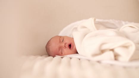 Cute-newborn-baby-boy-laying-on-soft-white-sofa-covered-with-blanket-slowly-falling-asleep
