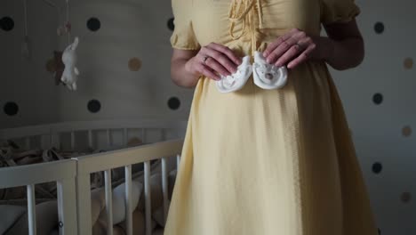 Pregnant-woman-in-yellow-dress-standing-next-to-wooden-crib-in-nursery-showing-cut-little-white-socks-for-the-baby