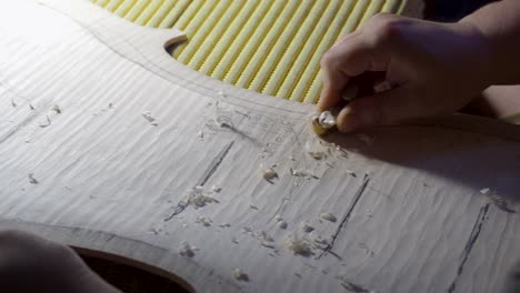 Stringed-instrument-maker-master-luthier-artisan-hand-proceed-with-graduation-work-on-cello-back-plate,-divide-wood-in-areas-to-carve-different-thickness-with-planes,-Wood-chips-and-curls-pile-up
