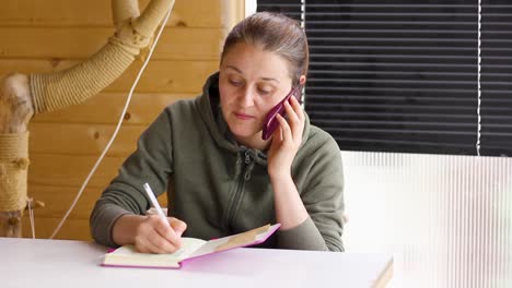 Woman-speaking-on-phone-while-looking-down-writing-in-notebook,-tilt-down