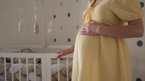 Pregnant-woman-in-yellow-dress-rubbing-her-belly-with-left-hand-while-standing-next-to-white-wooden-crib-in-nursery-with-dotted-wall-in-the-background