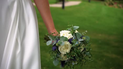 Close-up-shot-of-a-bride-walking-outdoor-in-the-park-holding-a-simple-bridal-bouquet-of-white-roses-and-greenery-with-blur-background