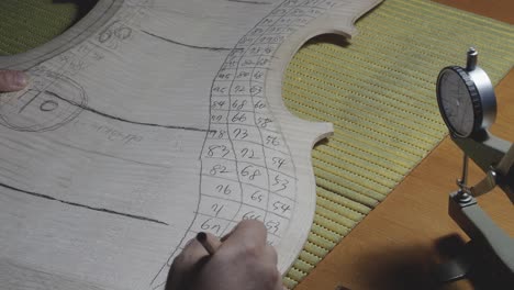 El-Maestro-Luthier-Fabricante-De-Instrumentos-De-Cuerda-Continúa-Con-El-Trabajo-De-Graduación-En-La-Placa-Posterior-Del-Violonchelo,-Usa-Un-Calibrador-De-Espesor,-Mide-Niveles,-Usa-Un-Lápiz-Para-Escribir-Valores-En-Madera,-Cremona,-Taller-De-Italia