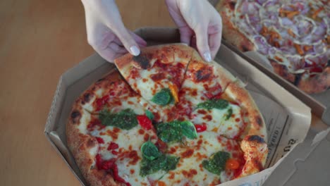 Eine-Handaufnahme-Von-Oben-Nach-Unten-Zeigt-Ein-Stück-Leckere-Neapolitanische-Pizza-Mit-Pesto-Sauce-In-Einem-Pizzakarton-Aus-Pappe-Auf-Dem-Tisch