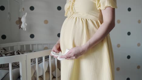 Pregnant-Woman-Standing-Next-To-Wooden-Crib-Holding-Baby-Sock-It-Her-Hands