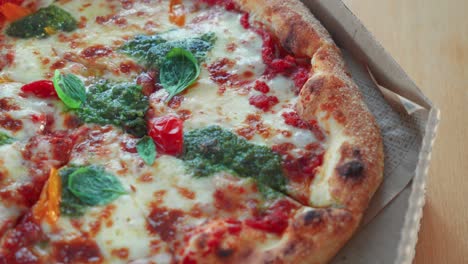 Closeup-shot-of-tasty-Neapolitan-pizza-with-pesto-sauce-in-a-cardboard-pizza-box-on-the-table,-rotating-shot
