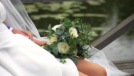 Close-up-shot-of-a-bride's-and-groom's-arms-holding-a-simple-bridal-bouquet-of-white-roses-and-greenery-with-a-background-of-lake-surface-in-the-rain
