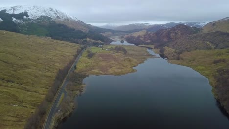 Aerial-flyover-fjord-and-road-in-scotland-highlands-during-grey-sky-and-snowy-mountains-in-Background
