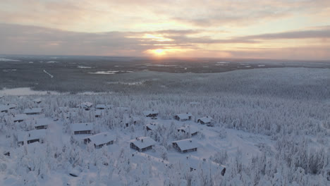 Drone-flying-over-a-mountaintop-cottage-town,-tilting-toward-the-sunset,-winter-in-Lapland