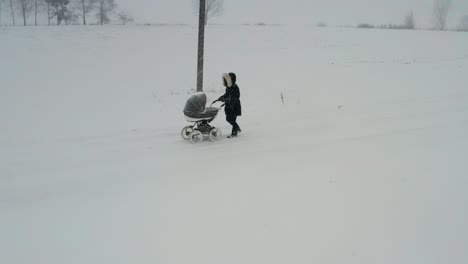 Female-in-black-winter-coat-walk-on-snowy-countryside-road-with-baby-carriage