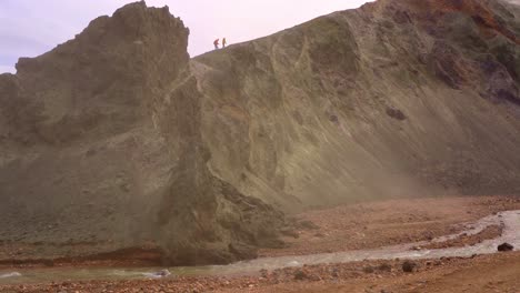 Silhouette-of-people-climbing-Bláhnúkur-mountain-in-Landmannalaugar,-Iceland,-with-a-river-and-green-cliff-in-the-foreground