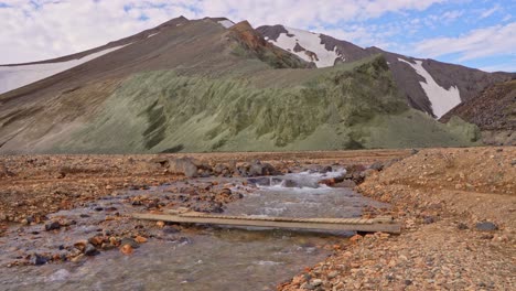 Fixed-wide-view-of-the-river-descending-from-Brennisteinsalda-in-Landmannalaugar,-Iceland,-featuring-a-small-wooden-bridge-for-hikers-in-front-of-Bláhnúkur-mountain