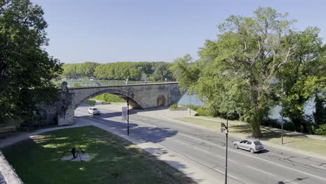 Street-by-the-river-under-the-historic-stone-bridge-in-Avignon-a-French-town-in-good-weather-with-few-cars
