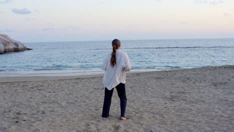 Young-girl-dancing-facing-peaceful-beach-at-sunset,-rear-wide-shot,-Tenerife