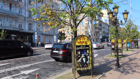 Schöne-Sonnige-Kiewer-Innenstadtstraße-Mit-Auf-Der-Straße-Fahrenden-Autos-Und-Wohnhäusern-In-Der-Ukraine,-Bäumen-Und-Lampen,-Gehenden-Menschen,-4K-Aufnahme