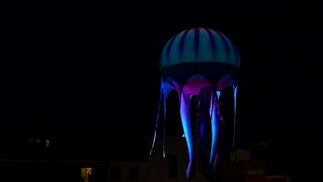 Blauer-Quallenballon,-Der-Nachts-Am-Dunklen-Himmel-über-Den-Häusern-Fliegt,-Während-Er-Beleuchtet-Wird