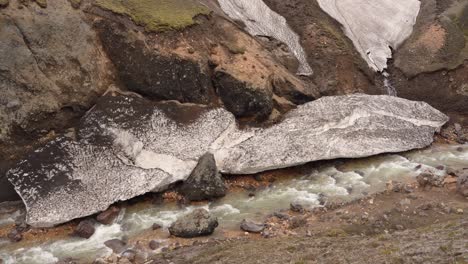 Fixed-view-of-the-river-descending-from-Brennisteinsalda-in-Landmannalaugar,-Iceland,-with-a-block-of-snow-along-the-river's-edge