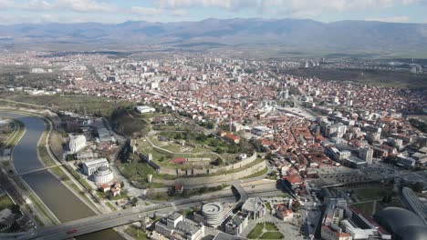 Drone-view-of-the-city-of-Skopje-crossing-the-Vardar-River,-stone-castle-in-the-city-center